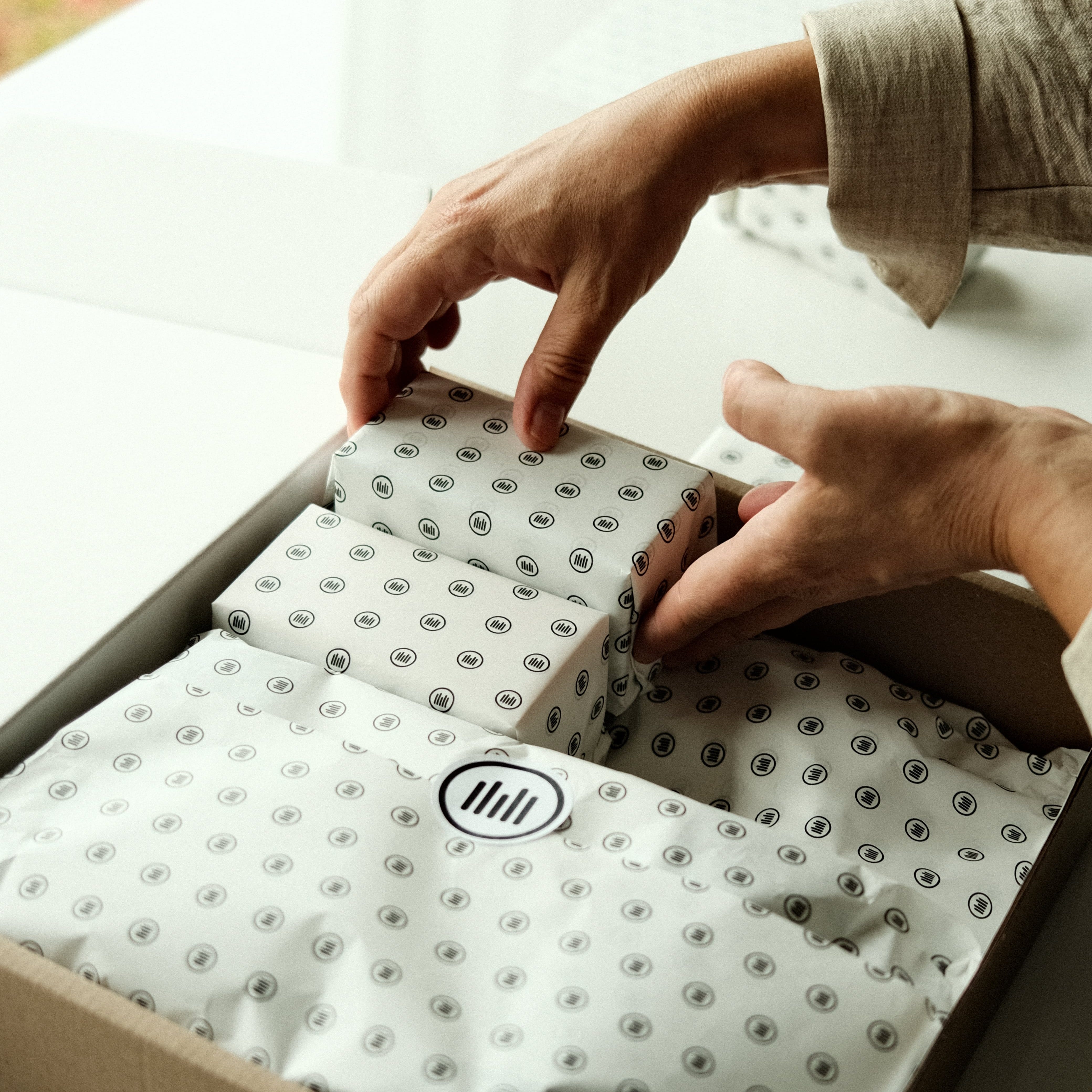 Hands unpacking a light brown box filled with white patterned packaging, revealing small items inside.