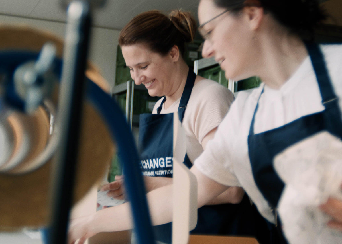 Two women in navy blue aprons working together, smiling in a vibrant, modern kitchen setting.